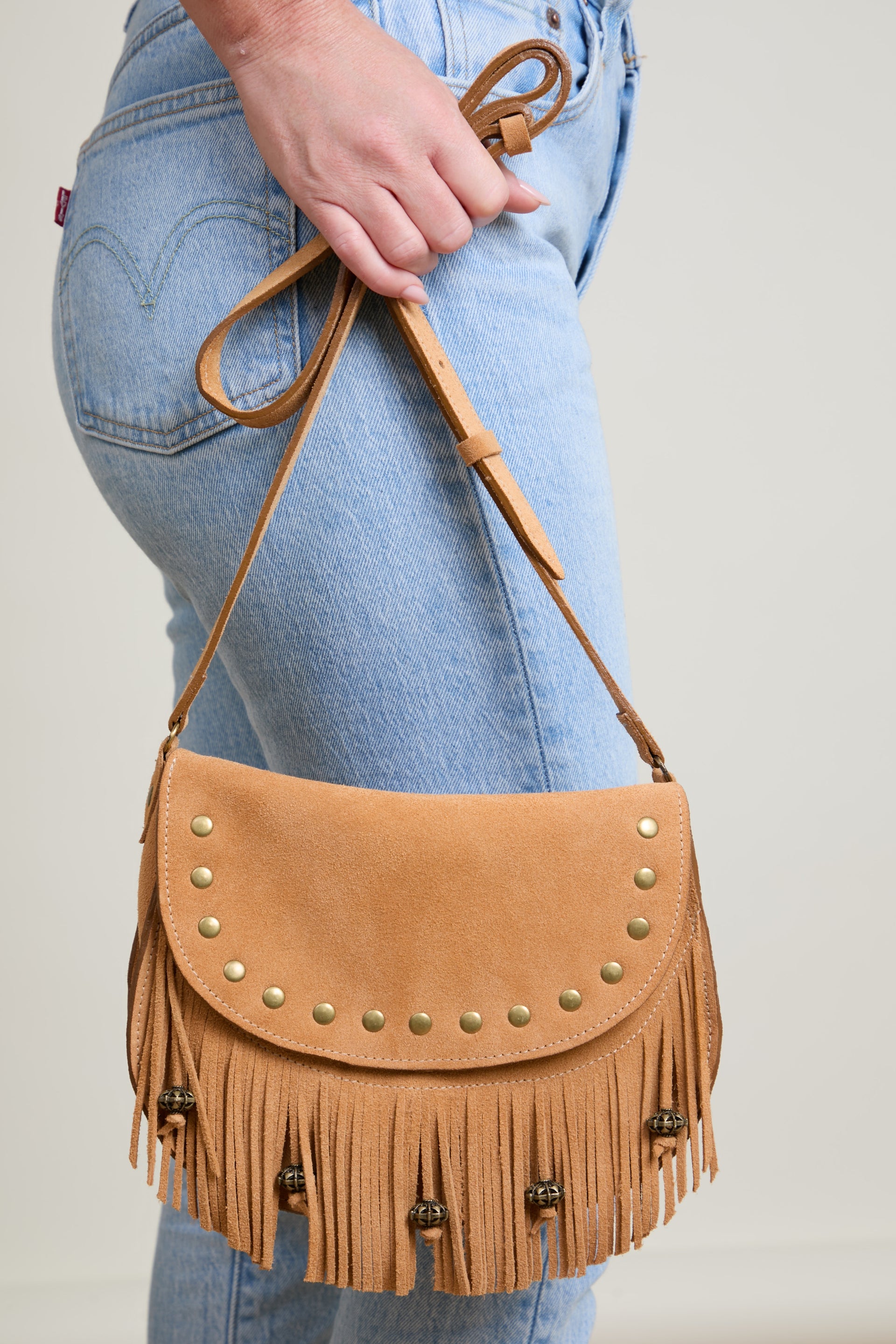 Tan leather handbag with fringe and studs held by a person wearing blue jeans.