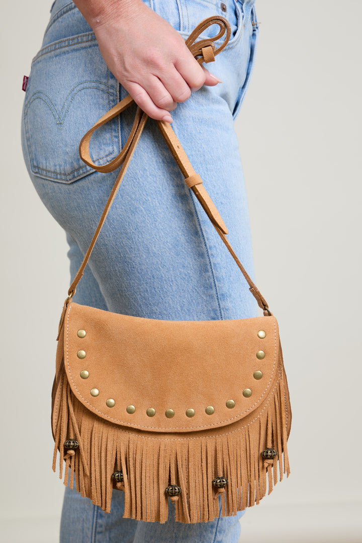 Tan leather handbag with fringe and studs held by a person wearing blue jeans.