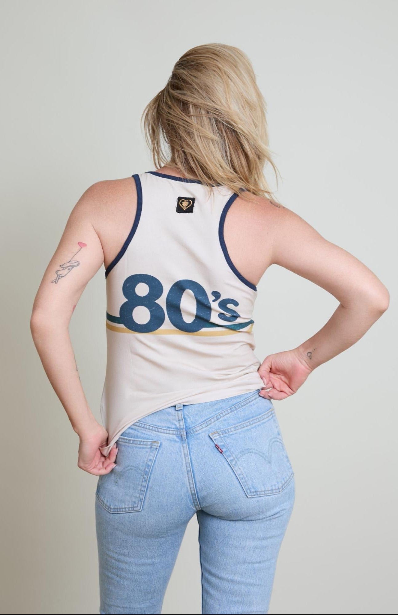 Person wearing a tank top with '80s' text and light blue jeans on a plain background