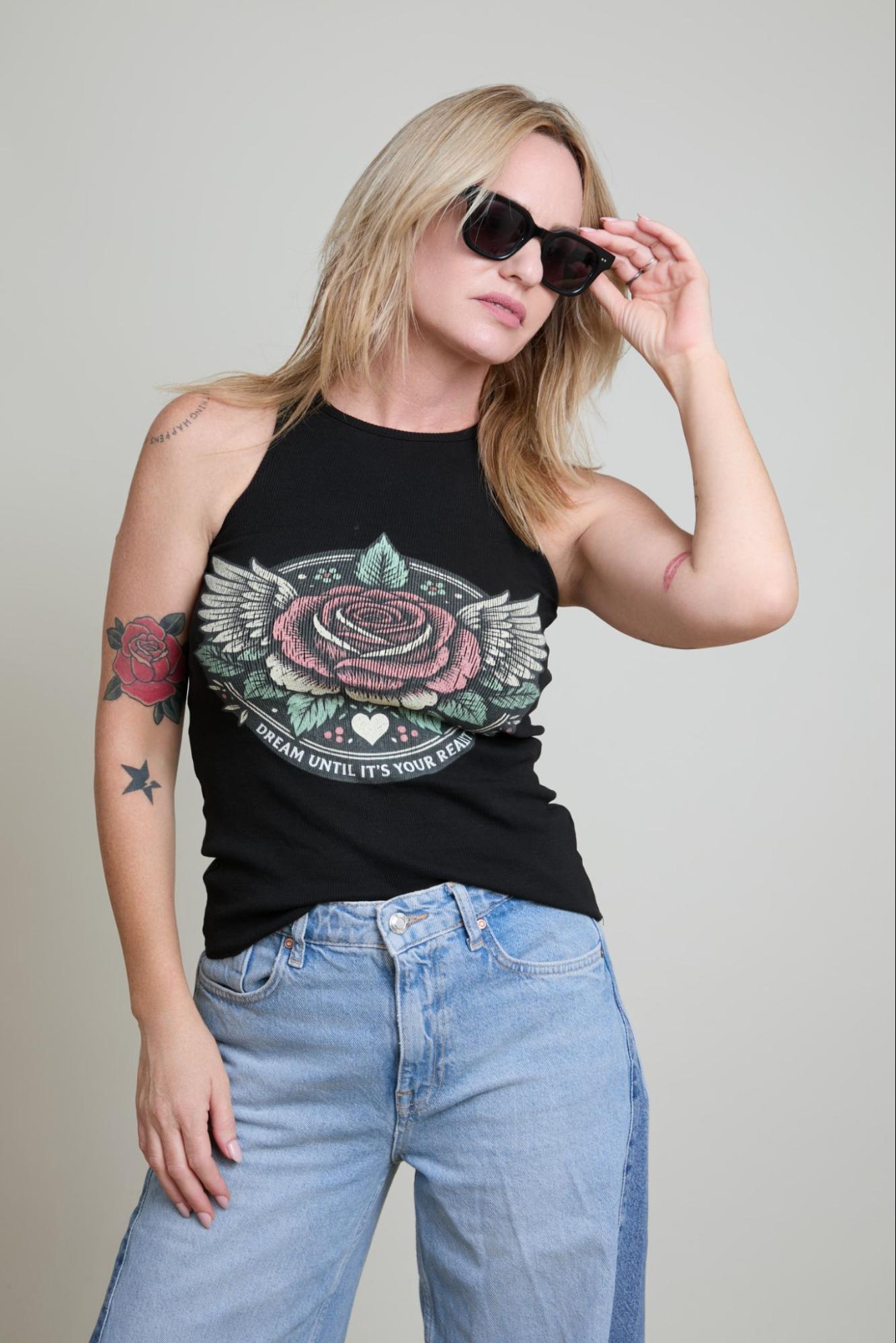 Woman wearing a black tank top with a colorful rose design and blue jeans, posing against a plain background.
