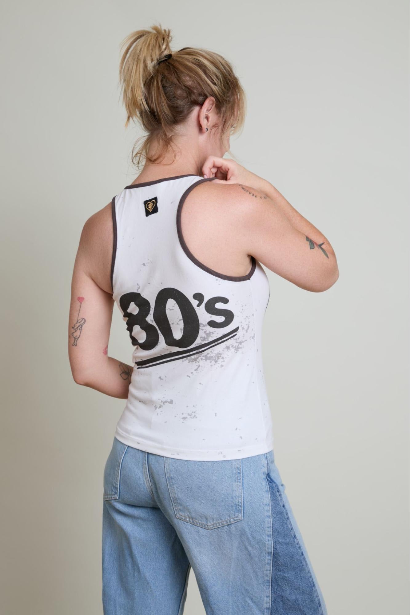 Person wearing a white tank top with '80's' text and blue jeans against a plain background