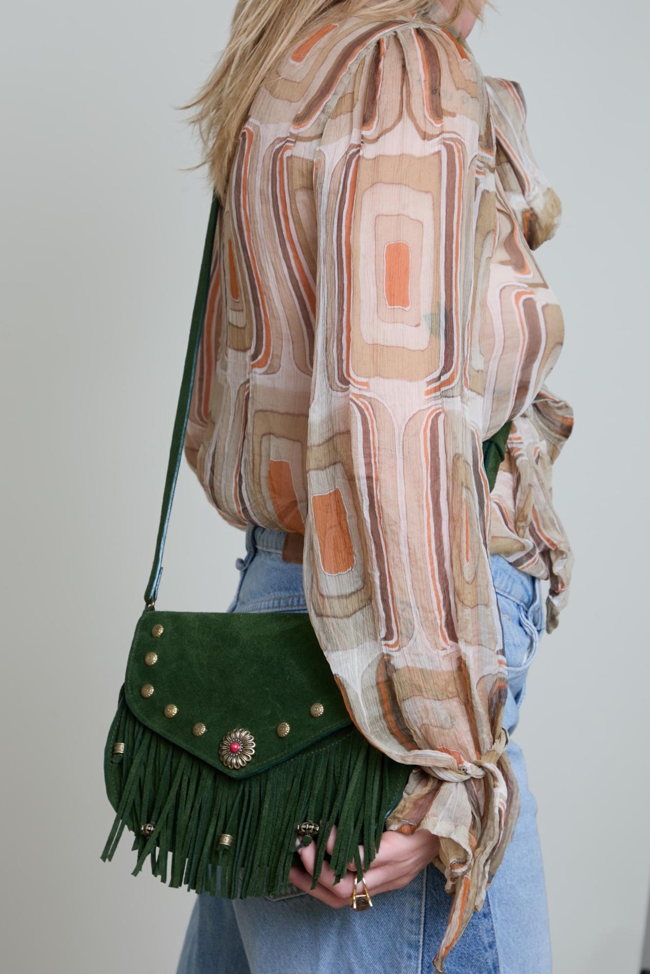 Person wearing a patterned scarf and holding a green fringed handbag against a plain background