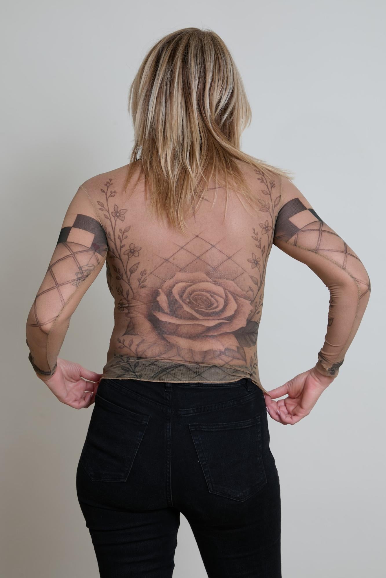 Person wearing a sheer top with tattoos and black pants against a plain background