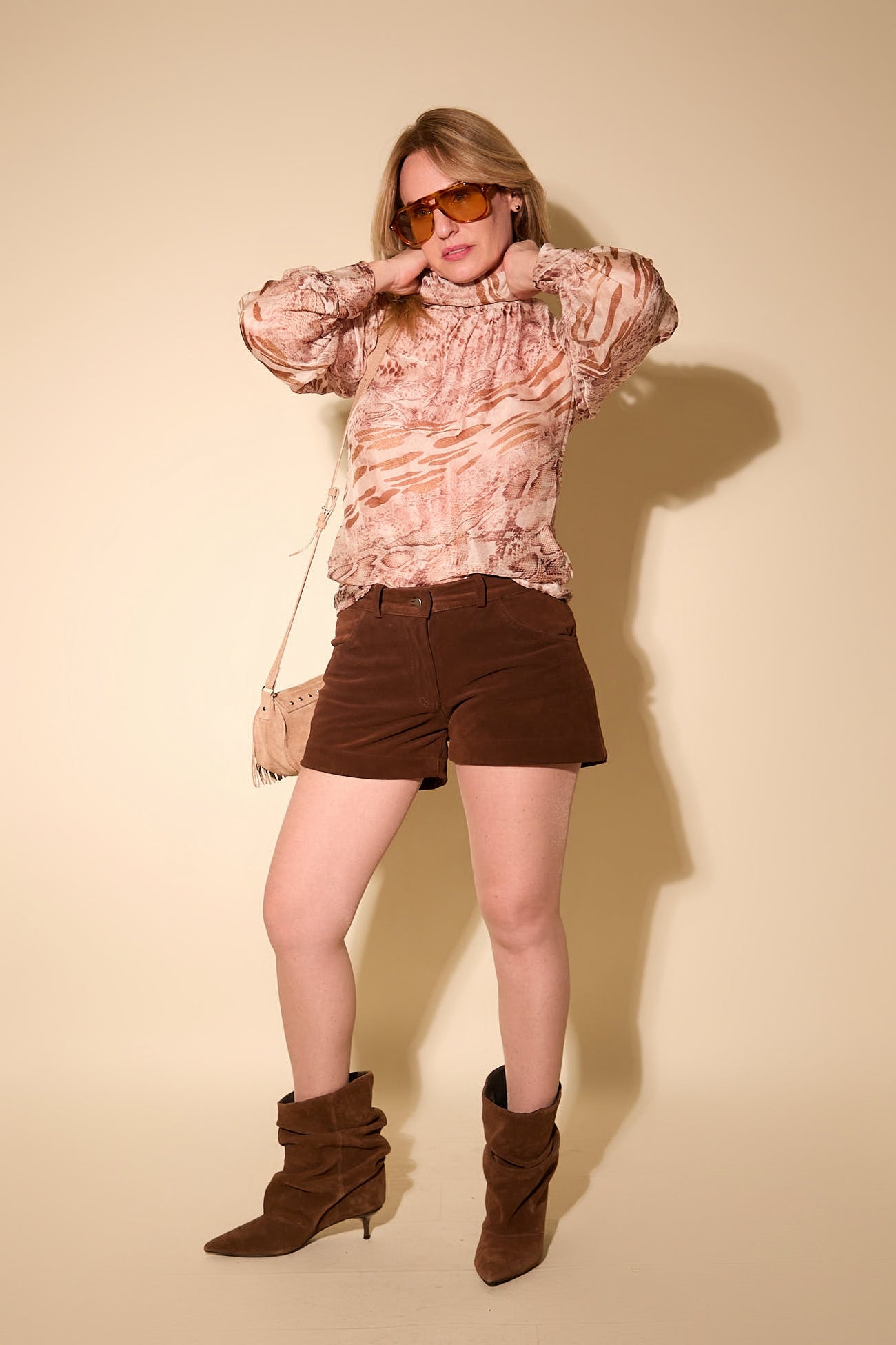 Woman wearing a patterned blouse, brown shorts, and brown boots against a beige background