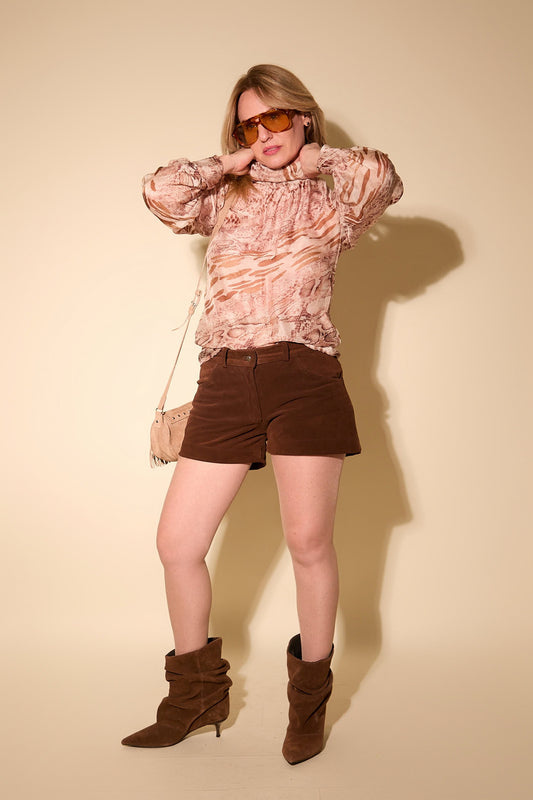 Woman wearing a patterned blouse, brown shorts, and brown boots against a beige background