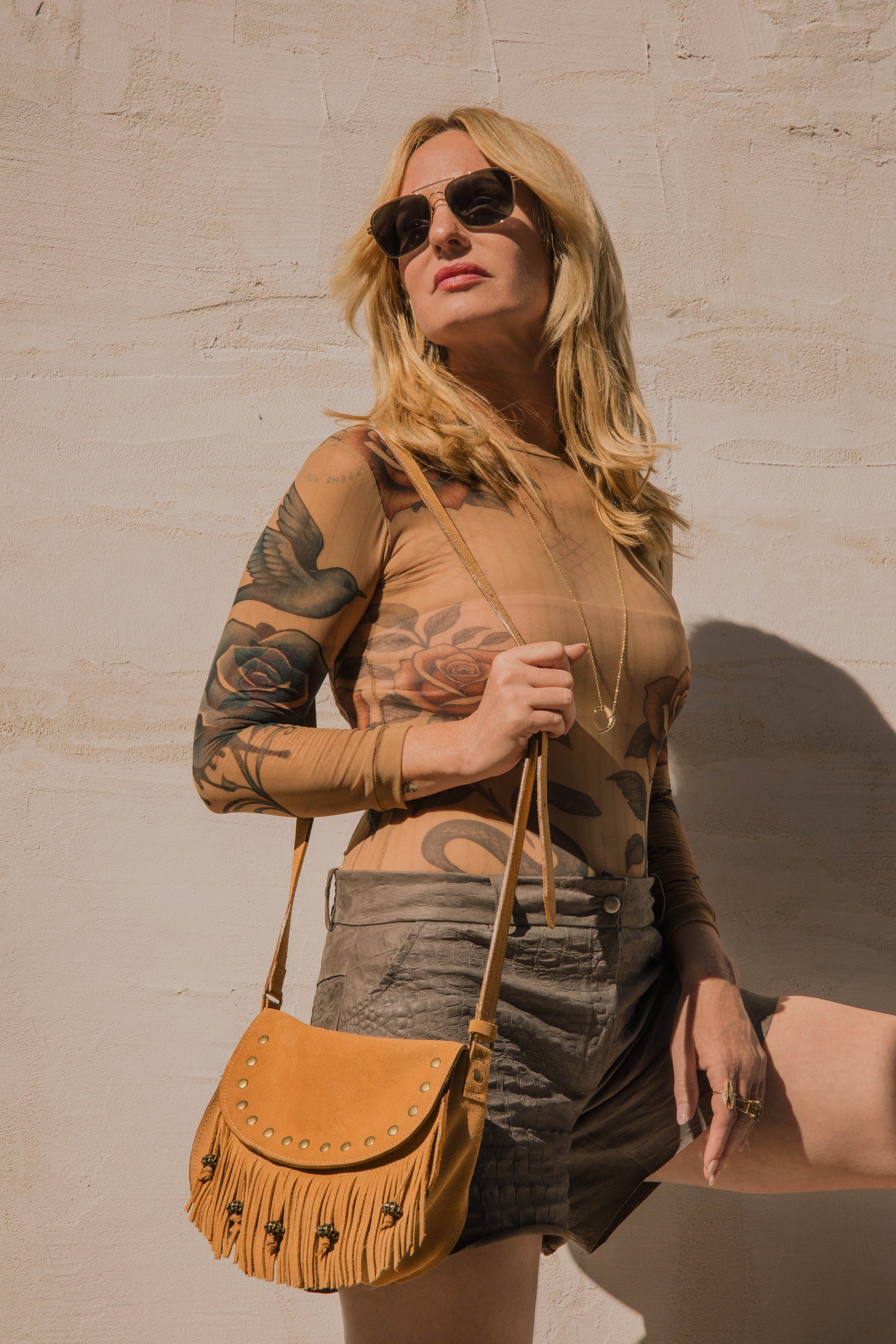 Woman with tattoos wearing sunglasses and a tan top, holding a brown leather bag against a beige wall.