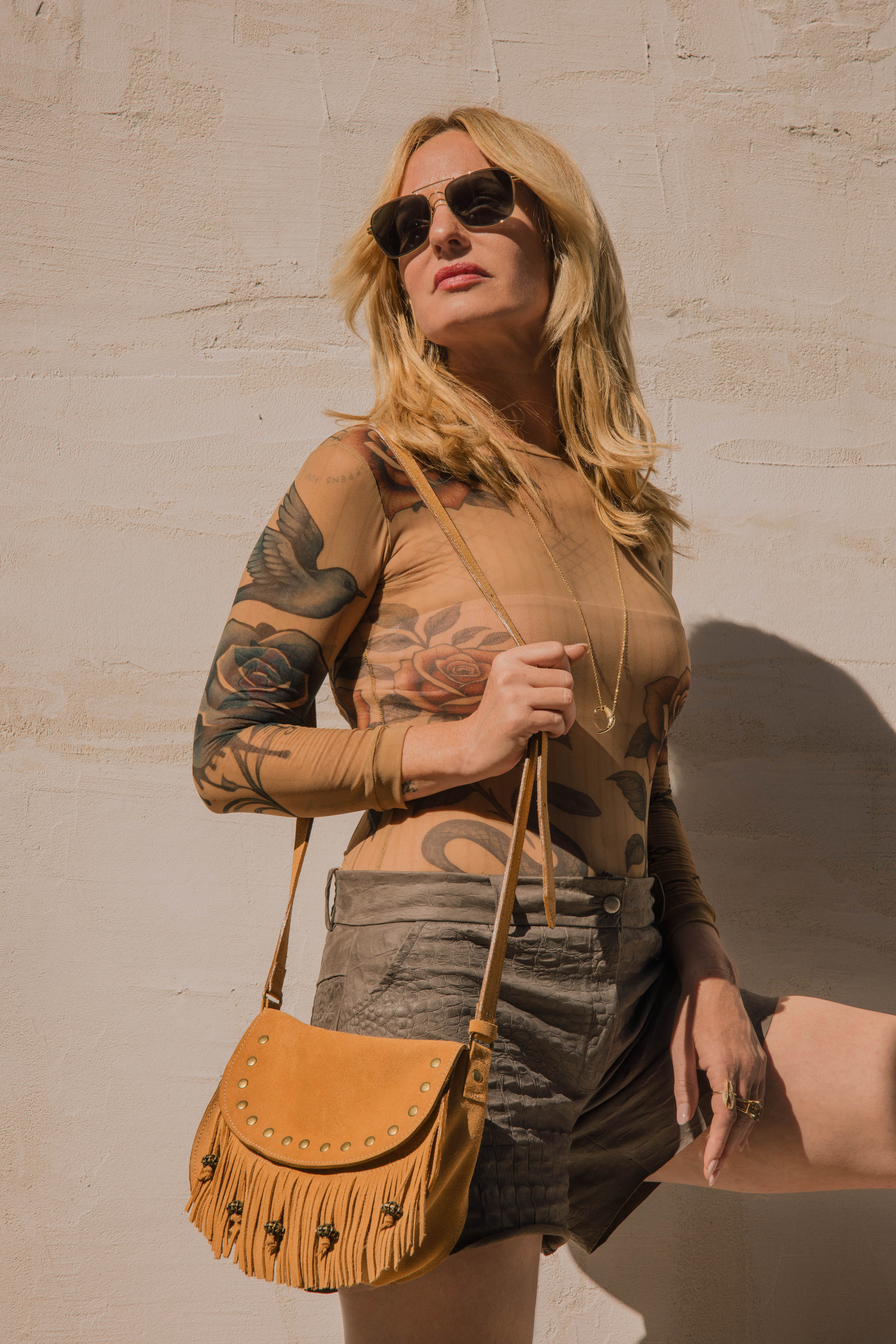 Woman with tattoos wearing sunglasses and a tan top, holding a brown leather bag against a beige wall.