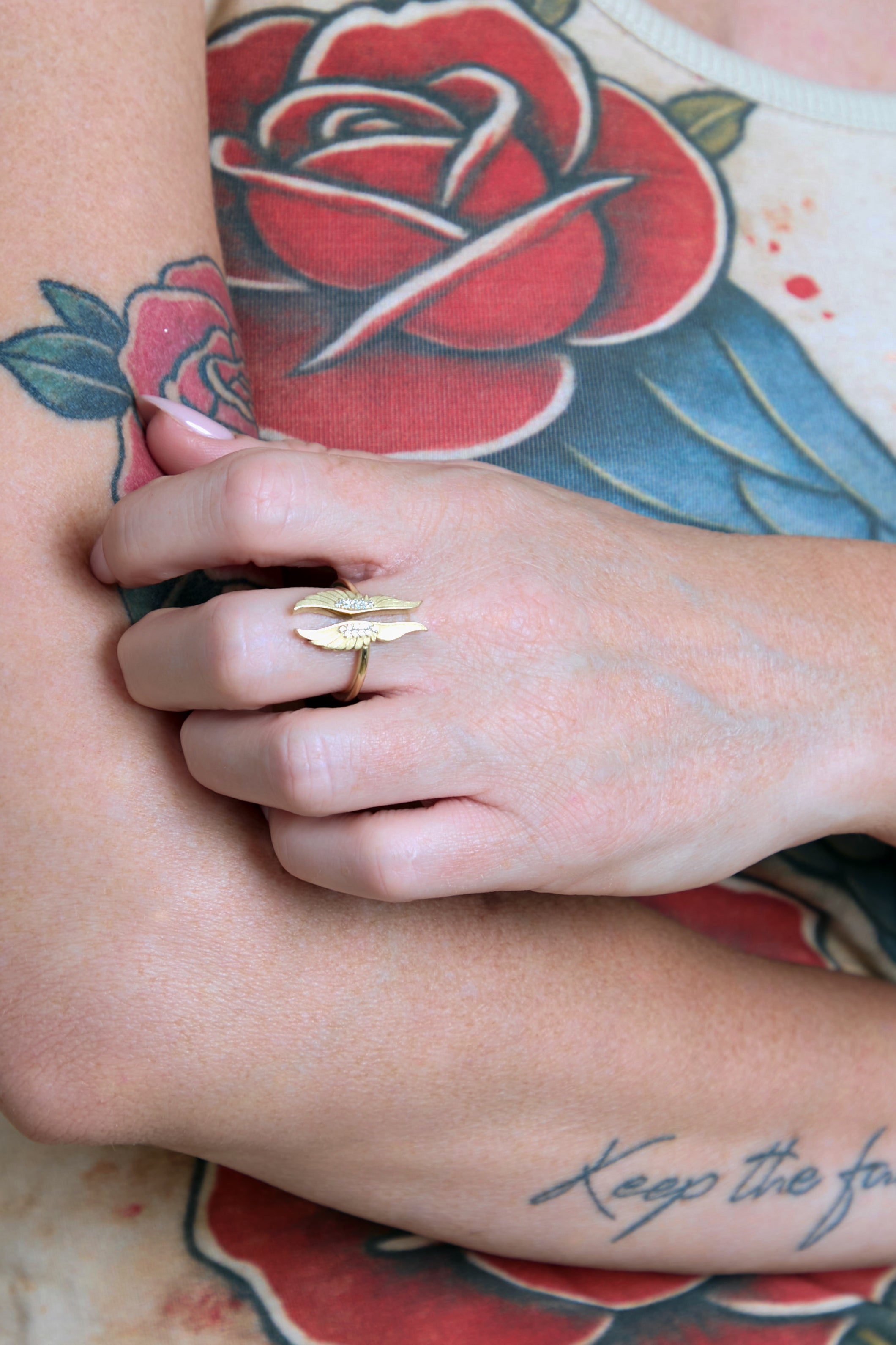 Hand wearing a gold ring with tattoos on arm and leg, featuring a red rose design.
