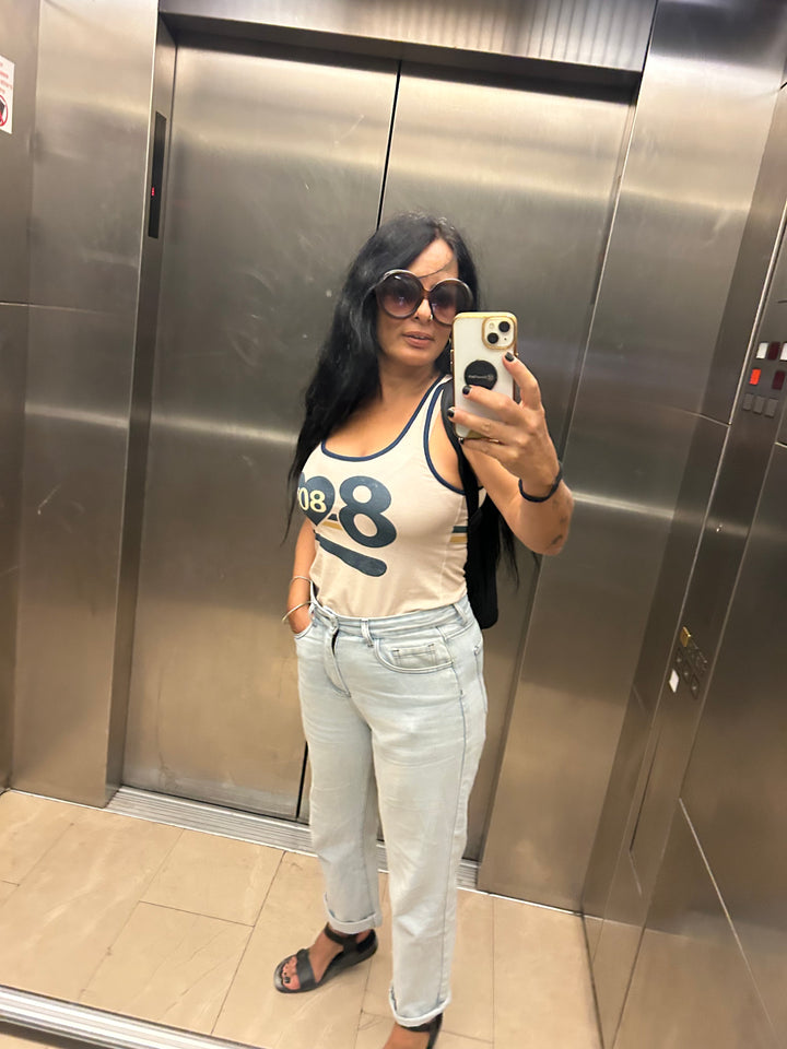 Person taking a mirror selfie in an elevator wearing a beige t-shirt with a number design and light blue jeans.
